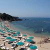 Spiaggia con sabbia chiara, ombrelloni turchesi e sdraio. Mare calmo e rocce sullo sfondo.