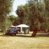 Campeggio con una tenda bianca, un'auto e alberi circostanti.