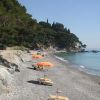 Una spiaggia di ciottoli con ombrelloni arancioni e vista sul mare. Alberi e rocce sullo sfondo.