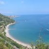 Spiaggia curva con mare blu e colline verdi. Una barca crea scie bianche nel mare.