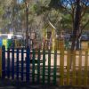 Parco giochi recintato con altalene e scivolo, circondato da alberi.