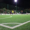 Campo da calcio notturno illuminato, con giocatori in azione e una rete sullo sfondo.