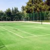 Campo da tennis all'aperto con erba sintetica verde e rete, circondato da alberi e recinzione.