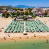 Spiaggia affollata con file di ombrelloni verdi, mare e case in lontananza.