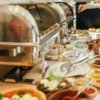 Buffet con piatti vari, persone che si servono cibo.