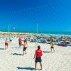Persone giocano a beach volley su sabbia bianca, mare con ombrelloni gialli sullo sfondo.