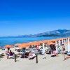 Spiaggia con file di ombrelloni arancioni, persone sulla sabbia, mare e montagne sullo sfondo.