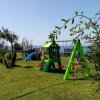 Area giochi per bambini su erba verde, con scivoli e altalene. Mare e alberi sullo sfondo.