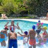 Bambini giocano in piscina, schizzando acqua, sotto la supervisione di un adulto in costume rosso.