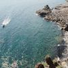 Una costa rocciosa con mare blu e una barca che lascia una scia sull'acqua.