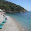 Spiaggia di sassi con lettini blu, mare calmo e colline verdi sullo sfondo.