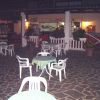 Cortile con tavoli e sedie in plastica, circondato da piante e un bar sullo sfondo.