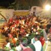 Un pubblico di persone sedute all'aperto, alcune in piedi, con un murale di sfondo.