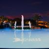 Piscina illuminata di notte con statue e palme.