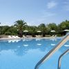 Piscina azzurra con palme e ombrelloni bianchi sullo sfondo, cielo sereno.