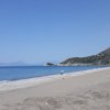 Spiaggia sabbiosa con mare calmo e colline sullo sfondo sotto un cielo azzurro.