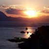 Tramonto sul mare con un'imbarcazione e montagne in lontananza, cielo arancione e nuvole.