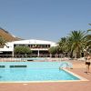 Piscina con persone che nuotano, palme sullo sfondo e un edificio bianco.