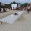 Una persona scivola su un telo in spiaggia, circondata da spettatori e coni colorati.