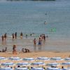 Persone in acqua poco profonda vicino alla riva, ombrelloni bianchi e blu sulla spiaggia sabbiosa.