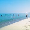 Persone che nuotano e camminano lungo una spiaggia con mare calmo e limpido sotto un cielo azzurro.