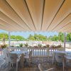 Tavoli e sedie bianche sotto una tenda, con vista su una piscina e giardino fiorito.