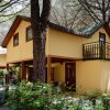 Una casetta gialla a due piani con balcone, circondata da alberi e vegetazione.