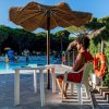 Uomo seduto sotto un ombrellone di paglia vicino a una piscina; alcuni bambini giocano in acqua.