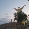 Statua con braccia aperte su collina al tramonto, persone camminano su sentiero roccioso.