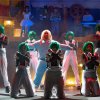 Persone con parrucche verdi e tute bianche eseguono una coreografia su un palco teatrale.