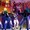 Performers con parrucche verdi ballano su un palco colorato, con due attori sullo sfondo.