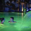 Due persone sorridenti in una piscina illuminata di verde con cascata d'acqua.