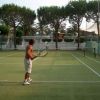 Una persona gioca a tennis su un campo all'aperto con alberi sullo sfondo.