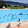 Piscina all'aperto con molte persone che nuotano, vista su un lago e colline.