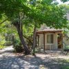 Piccola casa beige con veranda, immersa nel verde di alberi e arbusti.