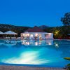 Una piscina illuminata di sera con vista su una struttura bianca e ampia vegetazione sullo sfondo.