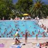 Ampia piscina esterna affollata di persone, con lettini e palme sullo sfondo.