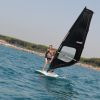 Persona che fa windsurf su acqua azzurra, vicino a una spiaggia affollata con pineta.