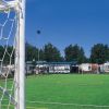 Campo da calcio in erba sintetica con rete in primo piano e camper dietro la recinzione.